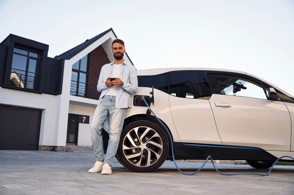 Beautiful young stylish man is with electric car at daytime charging the vehicle