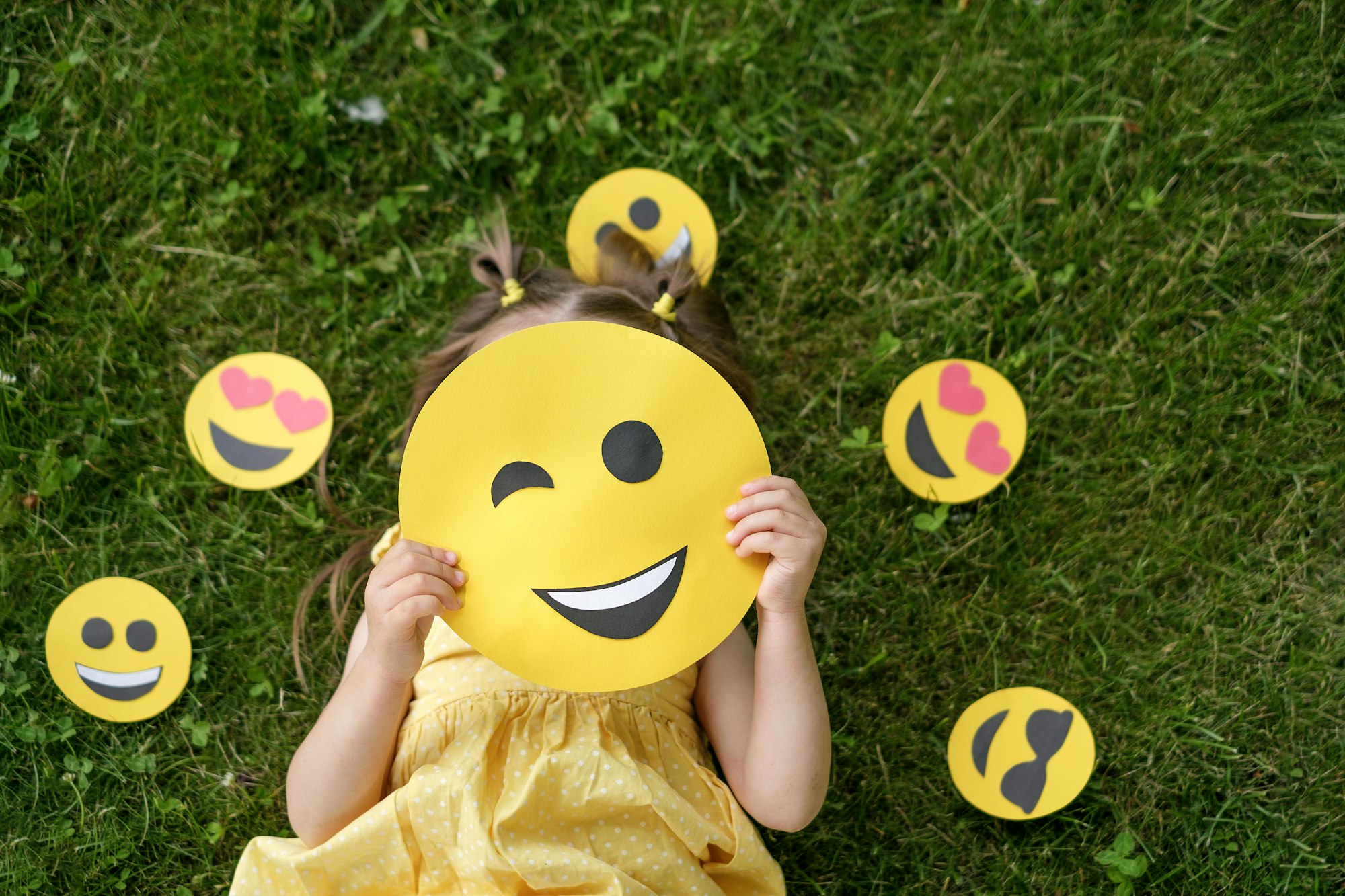 Kid hides his face behind a yellow winking emoticon lying on the grass in the park